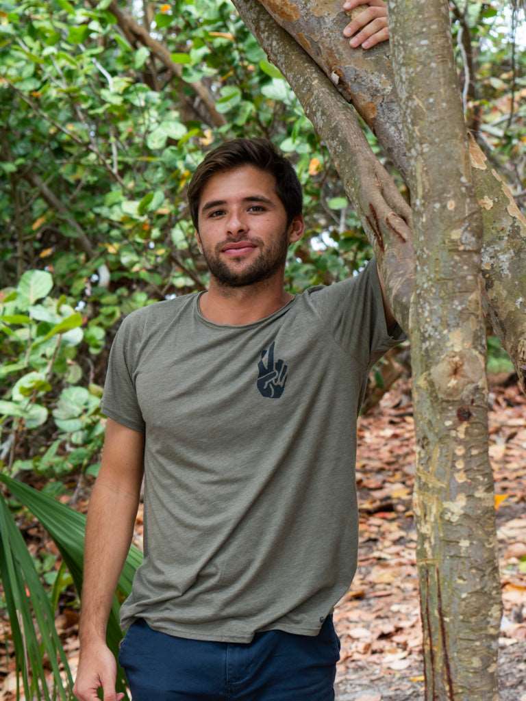 Man standing in a forest, wearing a green t-shirt with a logo, holding onto a tree.