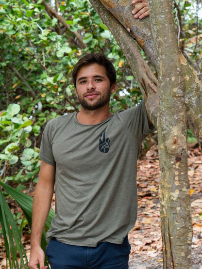 Man standing in a forest, wearing a green t-shirt with a logo, holding onto a tree.