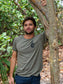 Man standing in a forest, wearing a green t-shirt with a logo, holding onto a tree.
