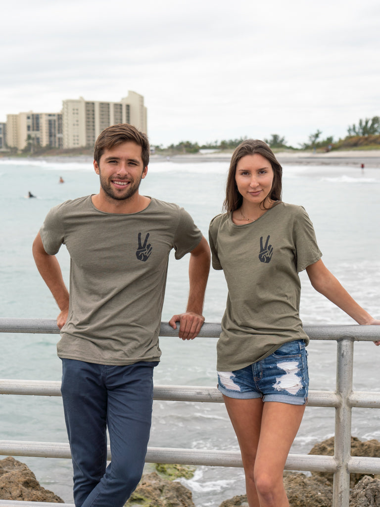 Man and woman standing on a bridge with the ocean in the background, wearing matching green t-shirts with a peace sign logo