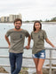 Man and woman standing on a bridge with the ocean in the background, wearing matching green t-shirts with a peace sign logo