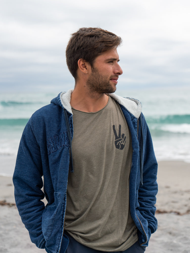 Man wearing blue hoodie with a green t-shirt underneath, featuring a peace sign graphic, standing on a beach