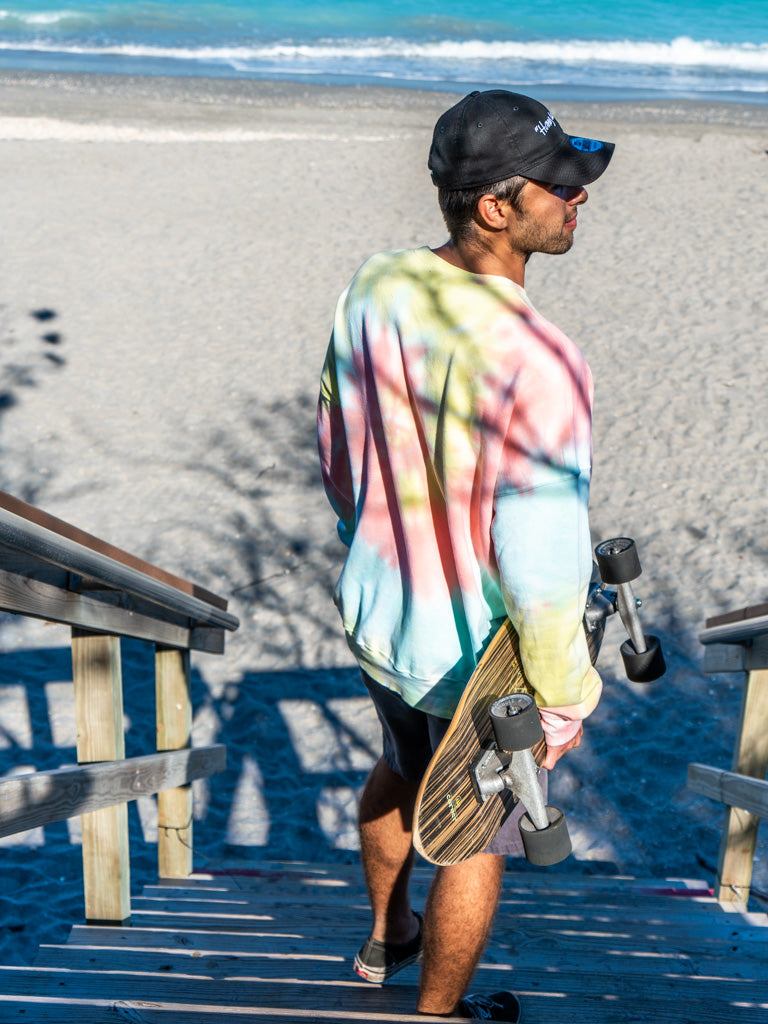 Man walking with a skateboard wearing a tie-dye sweatshirt with a peace symbol on a beach
