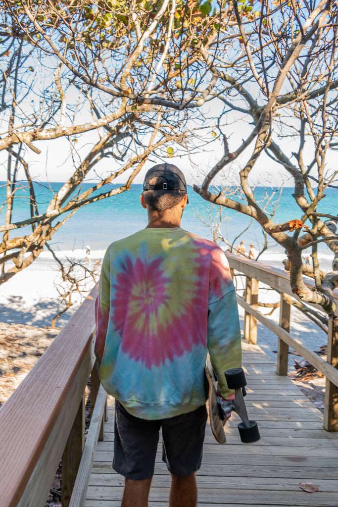 Man walking with a skateboard wearing a tie-dye sweatshirt with a peace symbol on a beach