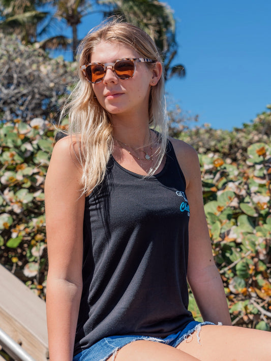 Woman wearing sunglasses and a black tank top with a logo, sitting outdoors.