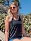 Woman wearing sunglasses and a black tank top with a logo, sitting outdoors.