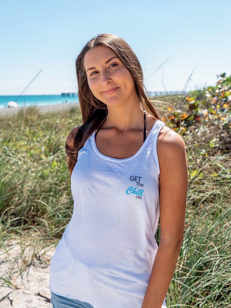 Woman wearing sunglasses and a white tank top with a logo, sitting outdoors.