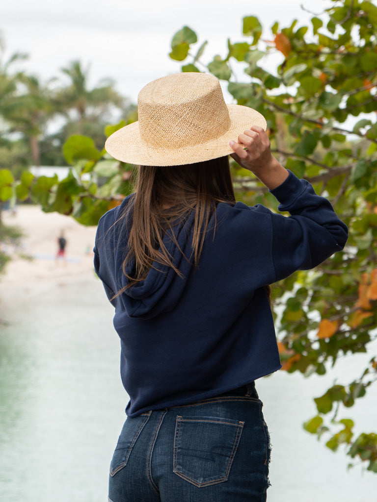 Woman wearing a navy hoodie, denim shorts, and a straw hat by a body of water.