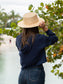 Woman wearing a navy hoodie, denim shorts, and a straw hat by a body of water.