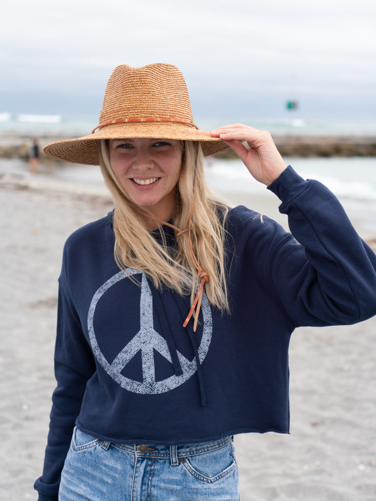 Woman wearing a navy hoodie with a peace symbol, denim shorts, and a straw hat by a body of water.