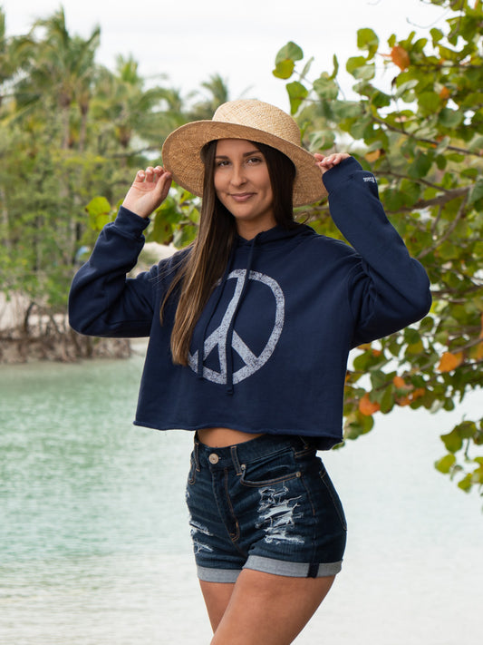 Woman wearing a navy hoodie with a peace symbol, denim shorts, and a straw hat by a body of water.