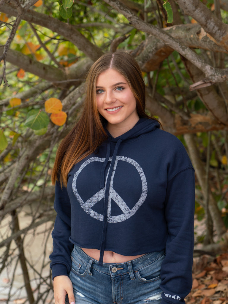 Woman wearing a navy hoodie with a peace symbol, and denim shorts by a body of water.