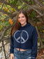 Woman wearing a navy hoodie with a peace symbol, and denim shorts by a body of water.