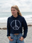 Woman wearing a navy hoodie with a peace symbol, denim shorts, and a straw hat by a body of water.