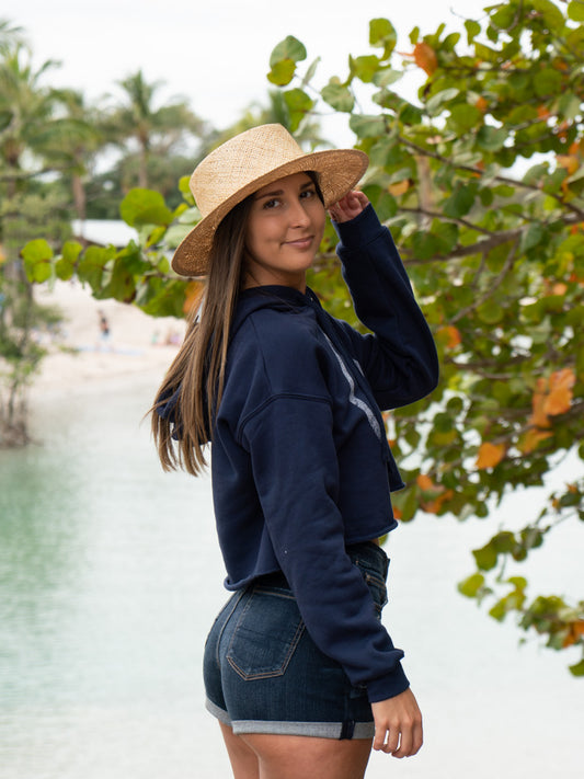 Woman wearing a navy hoodie with a peace symbol, denim shorts, and a straw hat by a body of water.