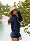 Woman wearing a navy hoodie with a peace symbol, denim shorts, and a straw hat by a body of water.