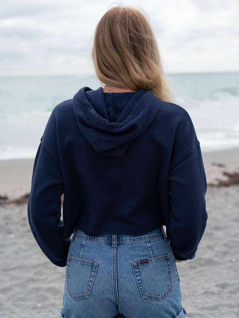 Woman wearing a navy hoodie, denim shorts, and a straw hat by a body of water.