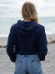 Woman wearing a navy hoodie, denim shorts, and a straw hat by a body of water.