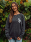 Woman wearing a dark gray sweatshirt with a peace sign design, standing in front of green foliage.
