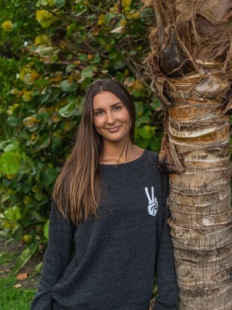 Woman wearing a dark gray sweatshirt with a peace sign design, standing in front of green foliage.