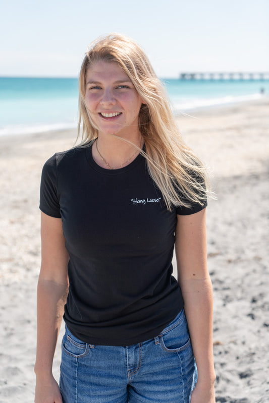 Woman wearing a black t-shirt with a logo, standing outdoors on a beach.