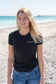 Woman wearing a black t-shirt with a logo, standing outdoors on a beach.