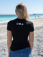 Woman wearing a black t-shirt with a logo, standing outdoors on a beach.