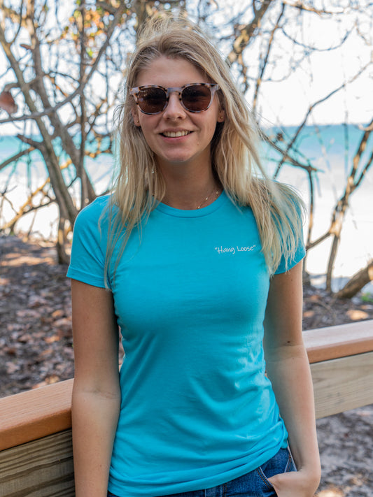 Woman wearing a turquoise t-shirt with a logo, standing outdoors with trees and a bridge in the background.