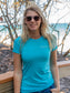Woman wearing a turquoise t-shirt with a logo, standing outdoors with trees and a bridge in the background.