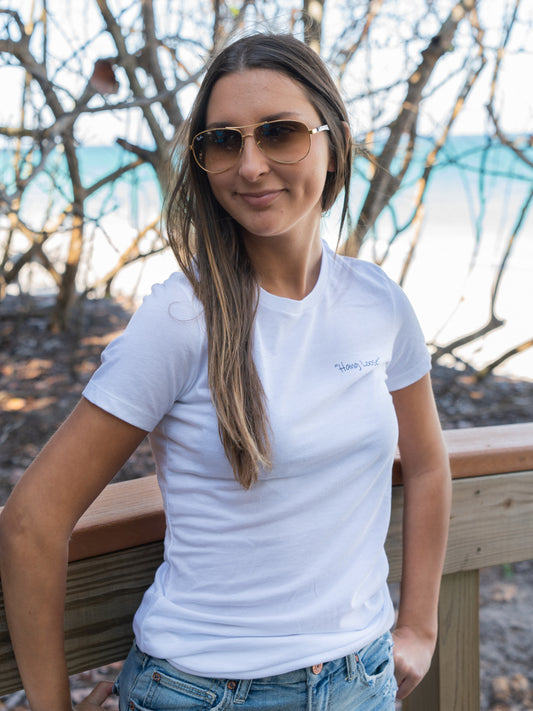Woman wearing a white t-shirt with a logo, standing outdoors with trees and a bridge in the background.
