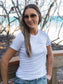 Woman wearing a white t-shirt with a logo, standing outdoors with trees and a bridge in the background.