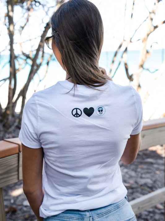 Woman wearing a white t-shirt with a logo, standing outdoors with trees and a bridge in the background.