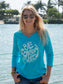 Woman wearing a turquoise long-sleeve shirt with a graphic design by a body of water.