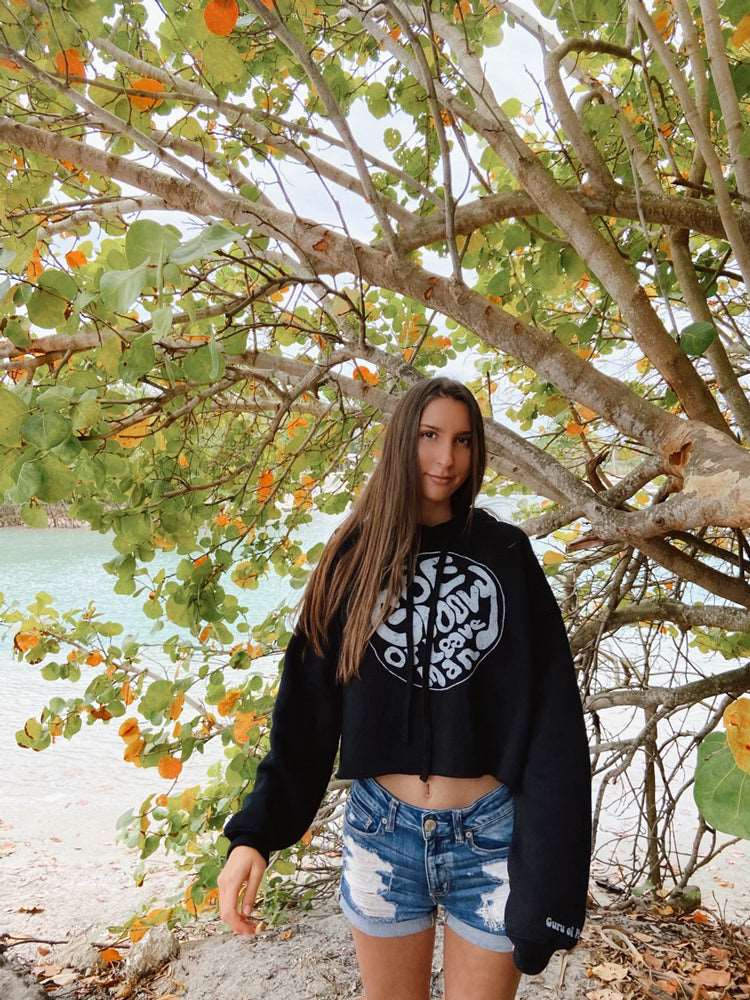 Woman wearing a black hoodie with white text on a beach with trees in the background