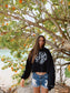 Woman wearing a black hoodie with white text on a beach with trees in the background