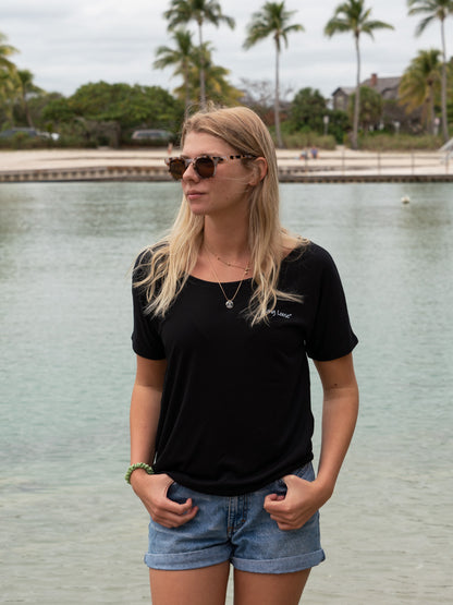 Woman in a black t-shirt and denim shorts standing by a body of water with trees in the background.