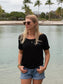 Woman in a black t-shirt and denim shorts standing by a body of water with trees in the background.