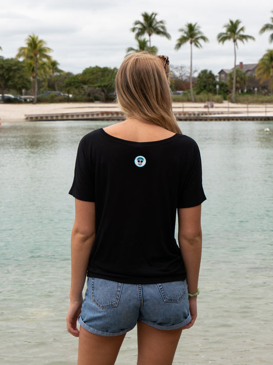 Woman in a black t-shirt and denim shorts standing by a body of water with trees in the background.