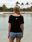 Woman in a black t-shirt and denim shorts standing by a body of water with trees in the background.