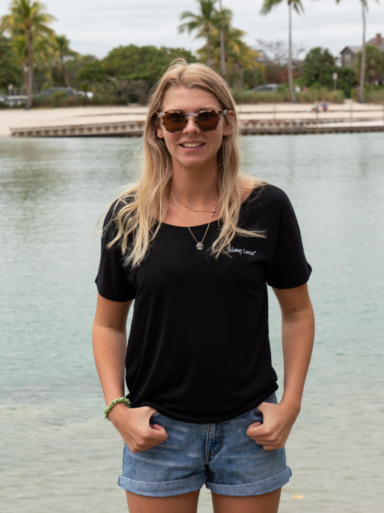 Woman wearing a black t-shirt and denim shorts standing by a body of water with trees in the background.