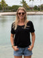 Woman wearing a black t-shirt and denim shorts standing by a body of water with trees in the background.