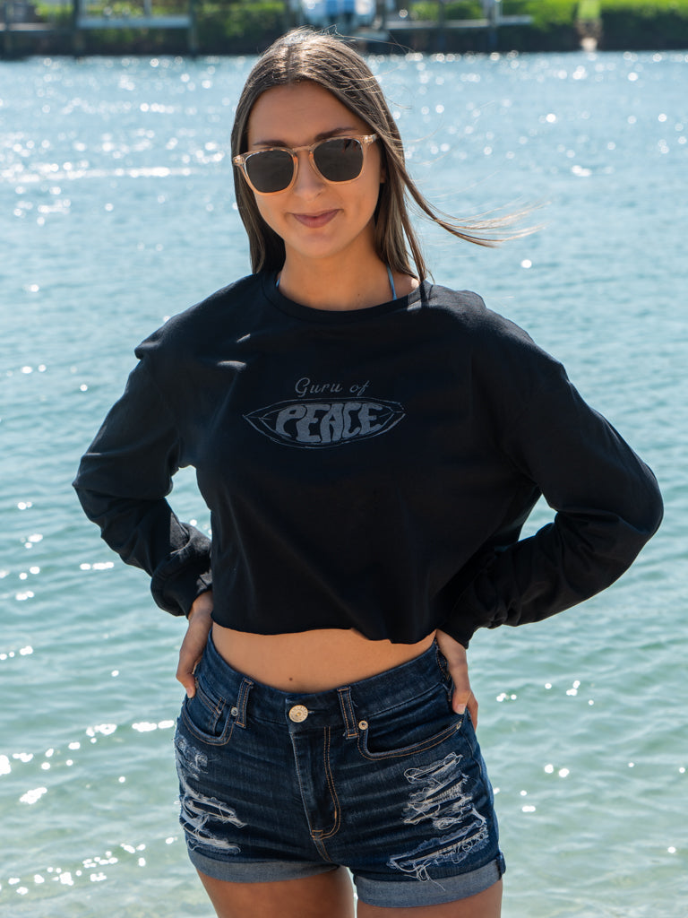Woman wearing a black long-sleeve crop top with a logo and text, standing by a body of water