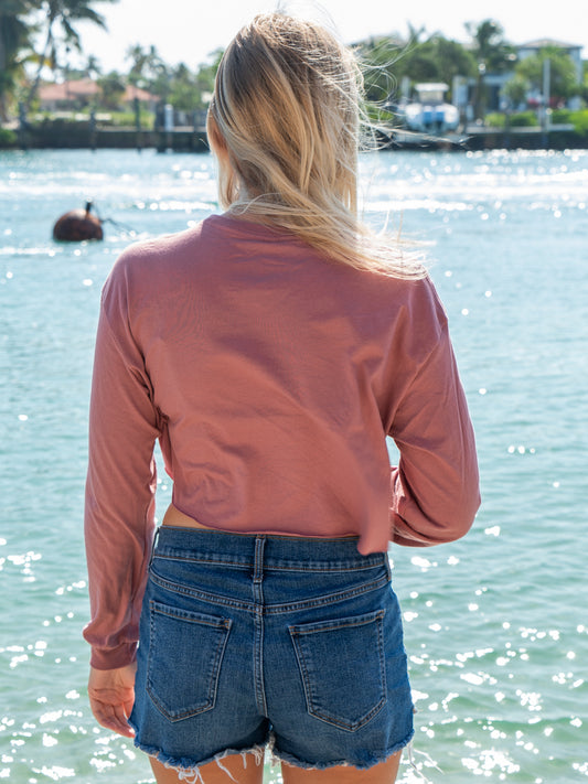 Woman wearing a mauve long-sleeve crop top with a logo and text, standing by a body of water