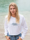 Woman wearing a white long-sleeve crop top with a logo and text, standing on a beach