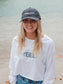 Woman wearing a hat and a white long-sleeve crop top with a logo and text, standing on a beach.