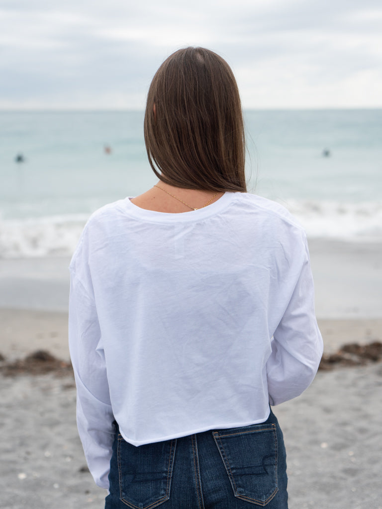 Woman wearing a white long-sleeve crop top with a logo and text, standing on a beach