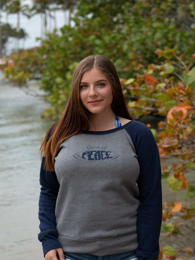 Woman wearing a gray and navy sweatshirt with 'Guru of Peace' text, standing outdoors by water.