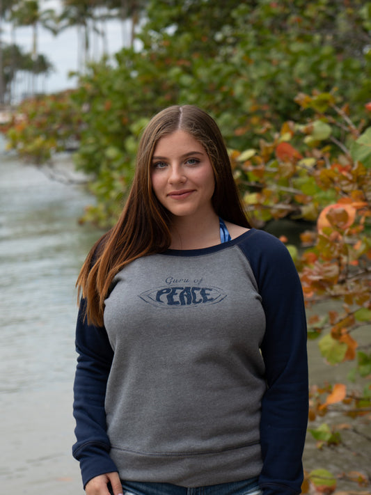 Woman wearing a gray and navy sweatshirt with 'Guru of Peace' text, standing outdoors by water.