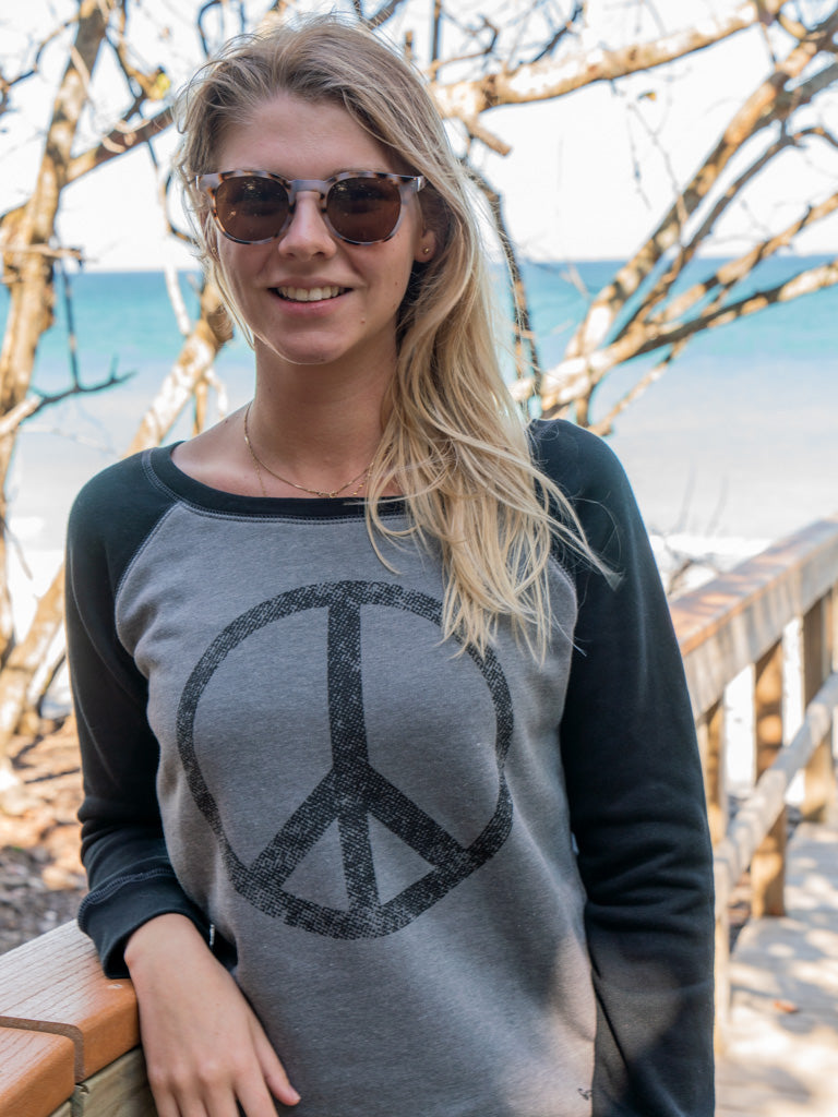 Woman leaning on a wooden railing wearing a crewneck featuring a large peace sign, surrounded by trees