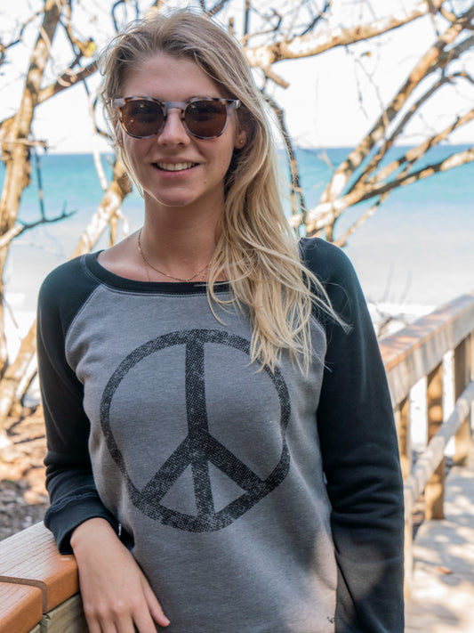 Woman leaning on a wooden railing wearing a crewneck featuring a large peace sign, surrounded by trees
